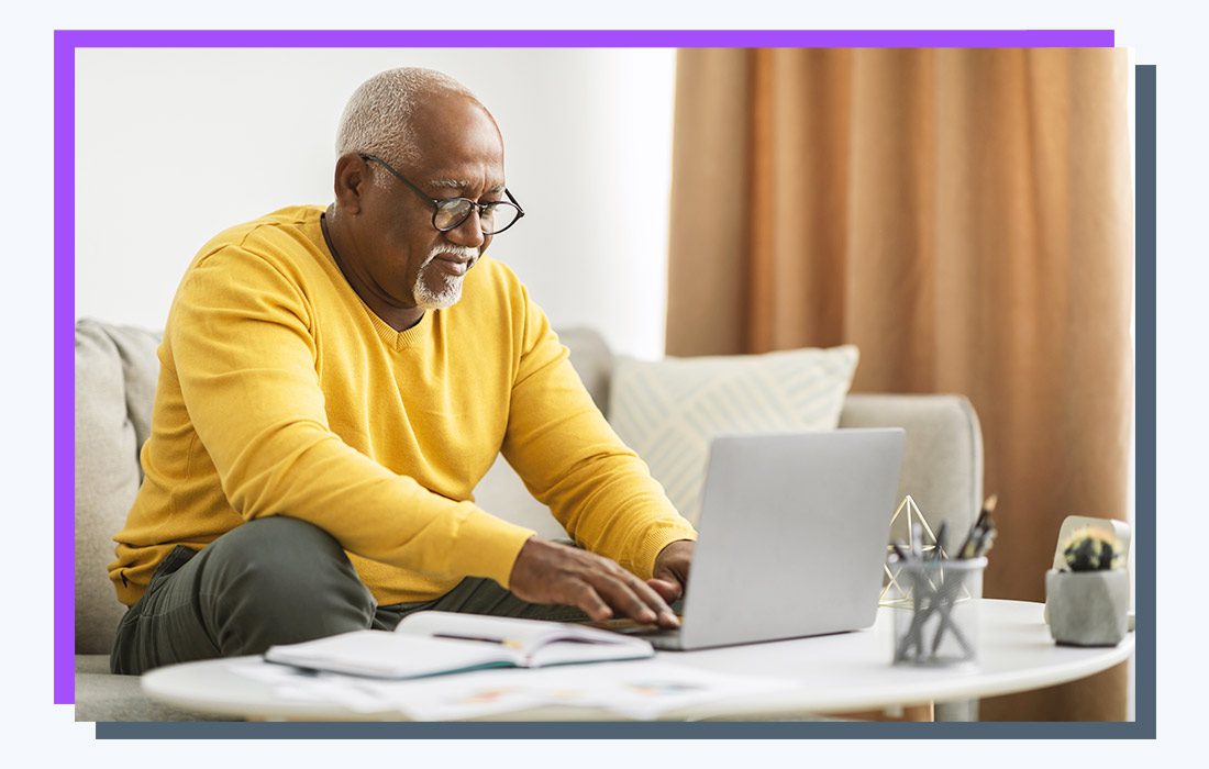 a man in a yellow sweater sitting on a couch and typing on a laptop.